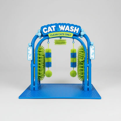 A cat walks through a blue and green self-cleaning cat grooming brush arch in a bright living room. The product has hanging green and blue brushes, a striped cat on the arch, and promotional text at the top reading 'Made for Daily Grooming' and 'CAT WASH CLEAN CATS ONLY'.